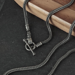 Puede incluir: Primer plano de un collar plateado con un diseño de cadena trenzada. El collar presenta un cierre único con una mano que sostiene una barra. La cadena se muestra sobre una superficie oscura con un fondo de madera.