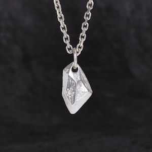 Könnte beinhalten: Halskette mit silberfarbenem Anhänger in geometrischer, facettierter Form. Der Anhänger hat ein kleines, kreisförmiges Muster. Die Kette ist ebenfalls silberfarben und hat ein Kettenglied-Design. Der Hintergrund ist einfarbig schwarz.
