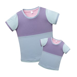 Könnte beinhalten: Zwei kurzärmelige T-Shirts, eines größer und eines kleiner, in einem passenden Farbschema. Die Shirts haben ein Colorblocking-Design mit Lavendel-, Hellblau- und rosa Akzenten. Die Ärmel sind hellblau und der Halsausschnitt ist rosa.