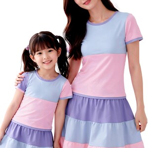 Könnte beinhalten: Zwei Personen in aufeinander abgestimmten Outfits in Pastellfarben. Die T-Shirts sind hellblau und rosa, mit lila Besatz. Die Röcke sind mit hellblauen, rosa und lila Bahnen versehen. Geeignet für Familienfotos. Die Outfits sind in einem Pastellfarbschema gehalten.