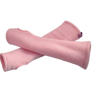 Puede incluir: Dos calentadores de brazo de forro polar rosa claro cruzados sobre un fondo blanco. Los calentadores de brazo tienen un aspecto suave y texturizado y están diseñados para cubrir el antebrazo y la mano, con una abertura para el pulgar.