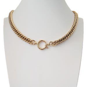 May include: A gold-coloured chain necklace with a thick, curb link design. The necklace features a circular clasp at the front. The jewellery is displayed on a white mannequin.