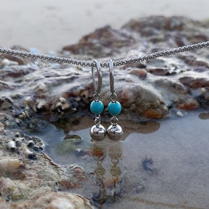 May include: A pair of silver hoop earrings with turquoise beads and silver ball charms. The earrings are hanging from a chain and are resting on a rocky surface.