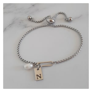 May include: Silver adjustable bracelet with a box chain. It has a rectangular charm with the letter "N" and a white pearl. The bracelet is shown on a white marble surface.