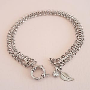 May include: A silver chain bracelet with three strands of different chain styles. The bracelet has a clasp closure and a small leaf charm with a clear crystal.