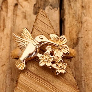 May include: A gold-toned ring featuring a detailed hummingbird in flight positioned next to a cluster of three small, five-petalled flowers. The metal surface has a polished finish that reflects light, highlighting the sculpted wings and floral details.