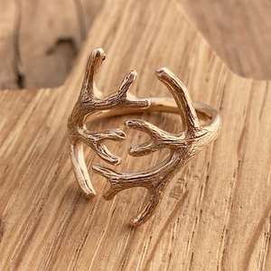 May include: Gold-toned ring featuring a design of two intertwined antlers. The ring is displayed on a wooden surface.