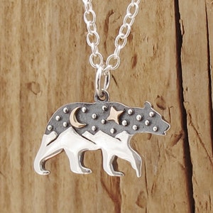 sterling silver bear pendant an oxidized top half of the hare has a raised bronze star and moon adding texture the bottom half of the bear has a polished mountain landscape the bear is walking in profile