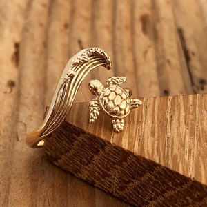 May include: Gold ring featuring a detailed turtle figurine riding a wave. The ring is made of metal and has a polished finish.