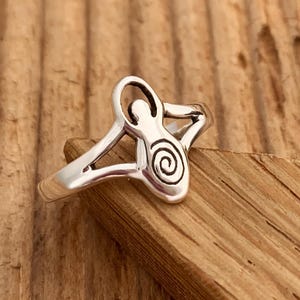 May include: A silver ring with a stylized female figure design. The figure is holding a spiral within her arms.