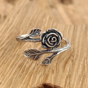 Könnte beinhalten: Ein silberner Ring mit einem Rosenmotiv. Die Rose ist um einen Zweig mit Blättern gewickelt.
