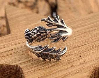 May include: A silver ring with a detailed design of a thistle flower. The ring is open and adjustable.