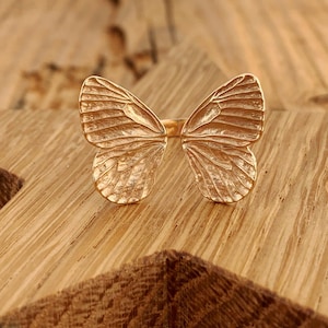 May include: A gold butterfly ring with detailed wings. The ring is on a wooden surface.