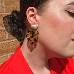 Puede incluir: Un par de pendientes de declaración dorados y marrones con un gran diseño de hoja tropical.