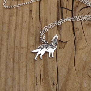 K&ouml;nnte beinhalten: Silberne Halskette mit einem Wolf-Anh&auml;nger. Der Wolf heult zum Mond und hat ein Bergketten-Design mit Sternen und einem Halbmond auf dem R&uuml;cken.