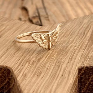 May include: A gold-colored ring with a detailed butterfly design. The ring features a delicate band and a prominent butterfly motif, showcasing intricate wing patterns. The ring is set against a wooden surface.