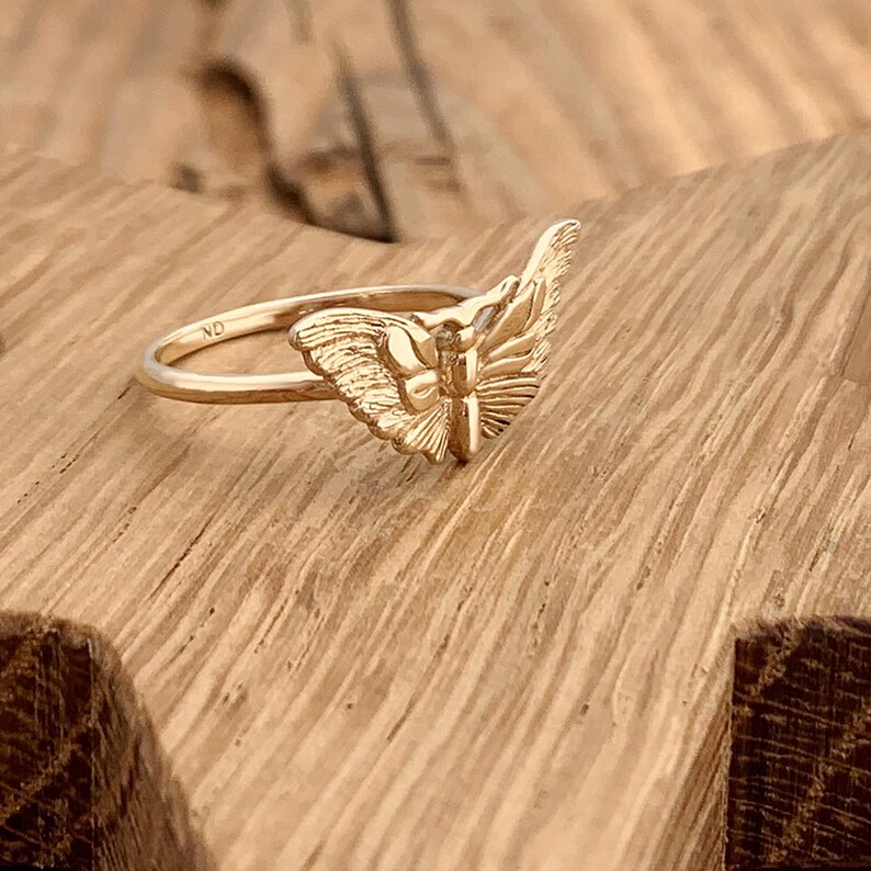 May include: A gold-colored ring with a detailed butterfly design. The ring features a delicate band and a prominent butterfly motif, showcasing intricate wing patterns. The ring is set against a wooden surface.