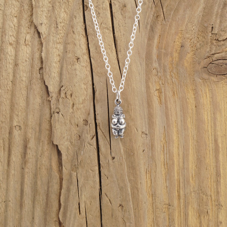 May include: Silver necklace with a small, detailed pendant depicting a female figure. The pendant is on a silver chain against a wooden background.