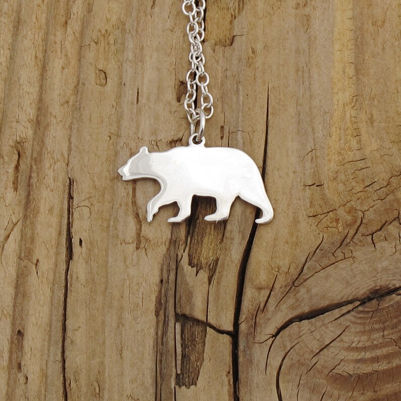 Pode incluir: Um colar com um pingente de urso de prata em um fundo de madeira. O urso &eacute; uma silhueta simples com um acabamento liso e polido.