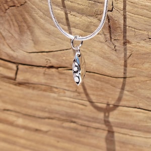 May include: A silver necklace with a pea pod pendant. The pendant has a polished, reflective surface with three white, pearl-like beads. The chain is a thin, textured silver snake chain. The background is a weathered, wooden surface.