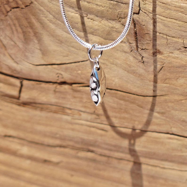 May include: A silver necklace with a pea pod pendant. The pendant has a polished, reflective surface with three white, pearl-like beads. The chain is a thin, textured silver snake chain. The background is a weathered, wooden surface.