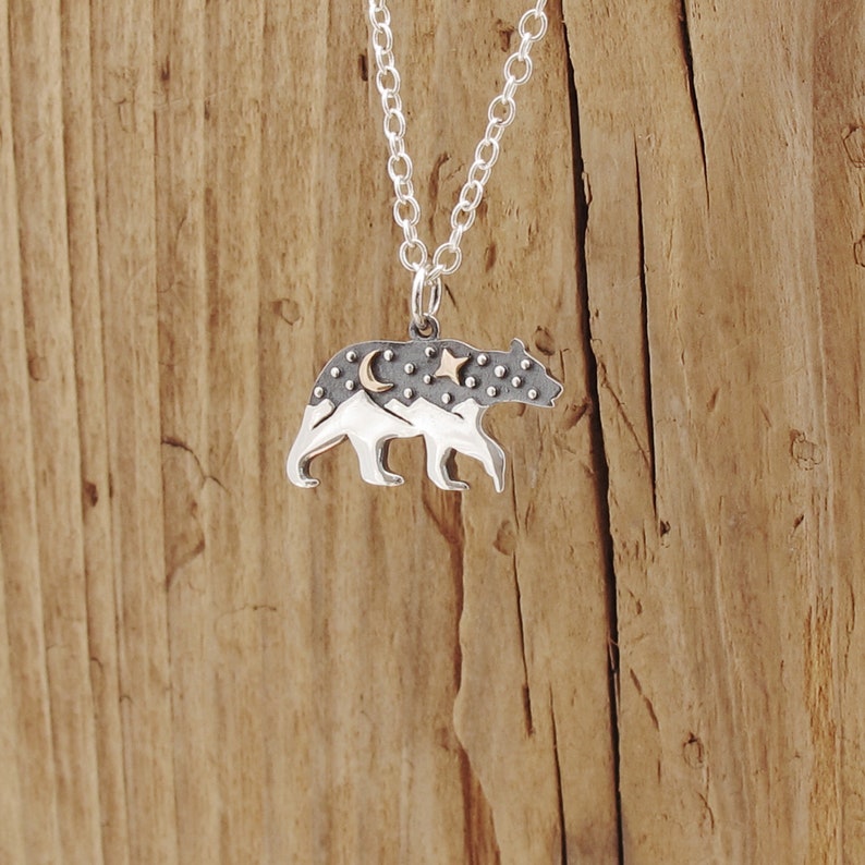 sterling silver bear pendant an oxidized top half of the hare has a raised bronze star and moon adding texture the bottom half of the bear has a polished mountain landscape the bear is walking in profile