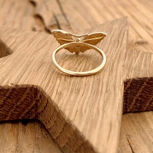 May include: A gold-colored ring with a butterfly design. The butterfly has detailed wing patterns and is centered on the ring's band. The ring is set on a wooden surface.