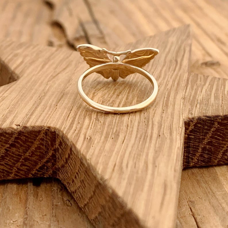 May include: A gold-colored ring with a butterfly design. The butterfly has detailed wing patterns and is centered on the ring's band. The ring is set on a wooden surface.