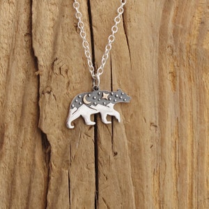 Pode incluir: Um colar de prata com um pingente de urso. O urso &eacute; feito de prata e tem um design de montanhas e lua em ouro nas costas. O urso est&aacute; em p&eacute; sobre uma superf&iacute;cie de madeira.