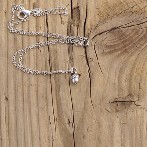 May include: A silver chain necklace with an acorn charm. The necklace is on a wooden surface.