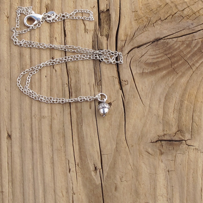 May include: A silver chain necklace with an acorn charm. The necklace is on a wooden surface.