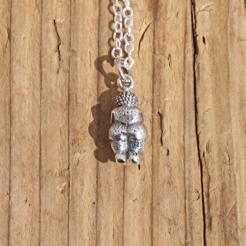May include: Silver necklace with a small, detailed pendant of a seated figure. The pendant is textured and has a small inscription on the back.