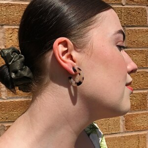 May include: A person wearing a pair of tortoiseshell hoop earrings. The earrings are a light brown colour with black speckles.