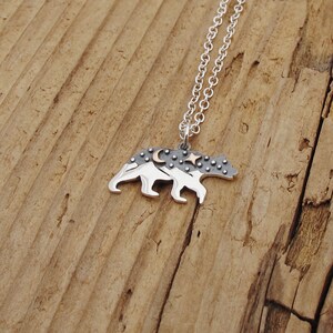 Pode incluir: Um colar de prata com um pingente de urso. O urso &eacute; contornado em prata e tem uma superf&iacute;cie texturizada com um design de lua e estrelas. O urso est&aacute; em p&eacute; sobre uma cordilheira.