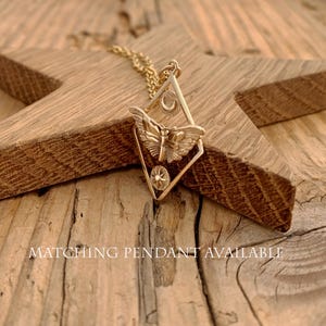 May include: A gold-colored pendant necklace featuring a butterfly design within a diamond-shaped frame. The frame includes a crescent moon and a starburst. The necklace rests on a wooden star. The text "MATCHING PENDANT AVAILABLE" is visible.