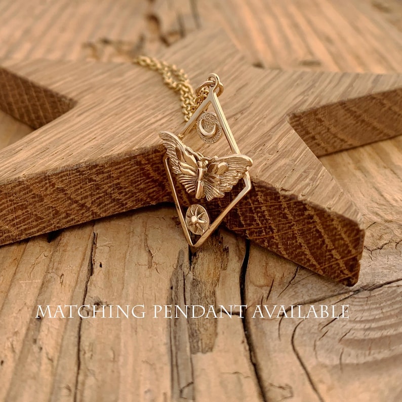 May include: A gold-colored pendant necklace featuring a butterfly design within a diamond-shaped frame. The frame includes a crescent moon and a starburst. The necklace rests on a wooden star. The text "MATCHING PENDANT AVAILABLE" is visible.