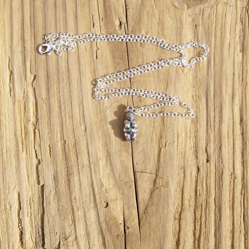 May include: A silver chain necklace with a small, detailed pendant in the shape of a person. The necklace is lying on a wooden surface.