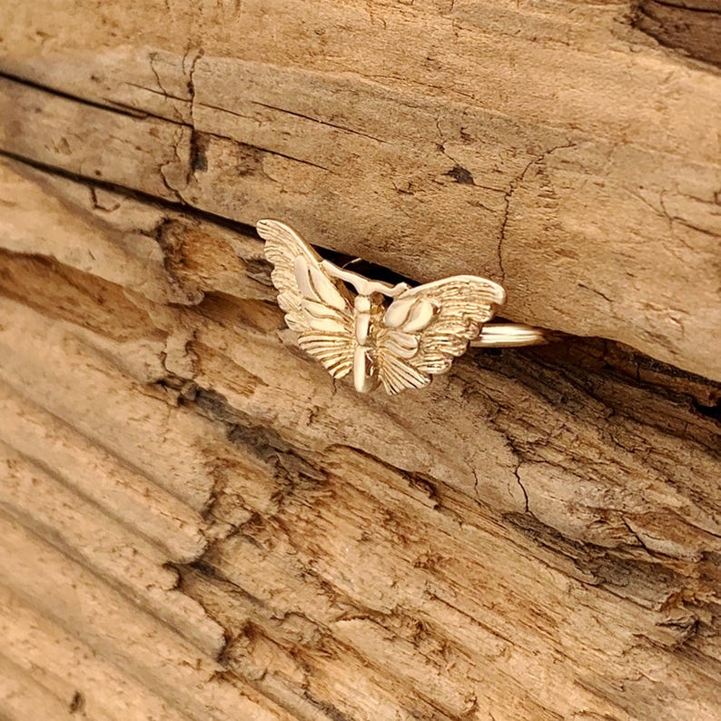 May include: A gold-colored butterfly ring. The ring features a detailed butterfly design with outstretched wings. The ring is set against a textured, wooden background.