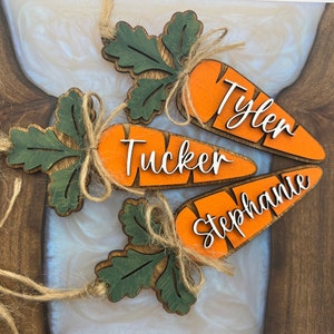 May include: Three wooden carrot decorations with green leaves and twine bows. Each carrot has a name written on it: Tyler, Tucker, and Stephanie.
