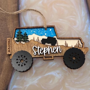 Peut inclure: Ornement de Noël en bois en forme de Jeep avec une scène de montagne enneigée et le nom Stephen. L'ornement est décoré d'un ciel bleu, de neige blanche et d'arbres verts. L'année 2024 est imprimée sur le côté de l'ornement.