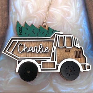 May include: A wooden dump truck ornament featuring a green Christmas tree on top with the year 2024 printed on it. The truck body is brown with white trim and black wheels. The name Charlie is written in white script on the side of the truck bed.