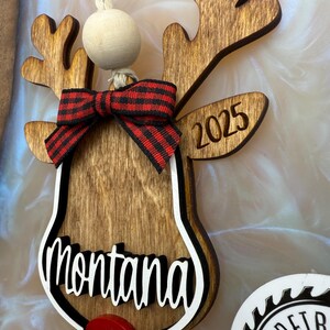 Puede incluir: Adorno de reno de madera con un lazo a cuadros rojo y negro, el nombre "montana" y el a&ntilde;o "2025" grabados en &eacute;l.