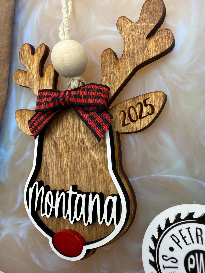 Puede incluir: Adorno de reno de madera con un lazo a cuadros rojo y negro, el nombre "montana" y el a&ntilde;o "2025" grabados en &eacute;l.