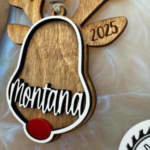 Puede incluir: Adorno de reno de madera con forma de campana y el nombre "Montana" escrito en &eacute;l. El adorno est&aacute; decorado con una nariz roja y el a&ntilde;o "2025" est&aacute; grabado en las astas del reno.