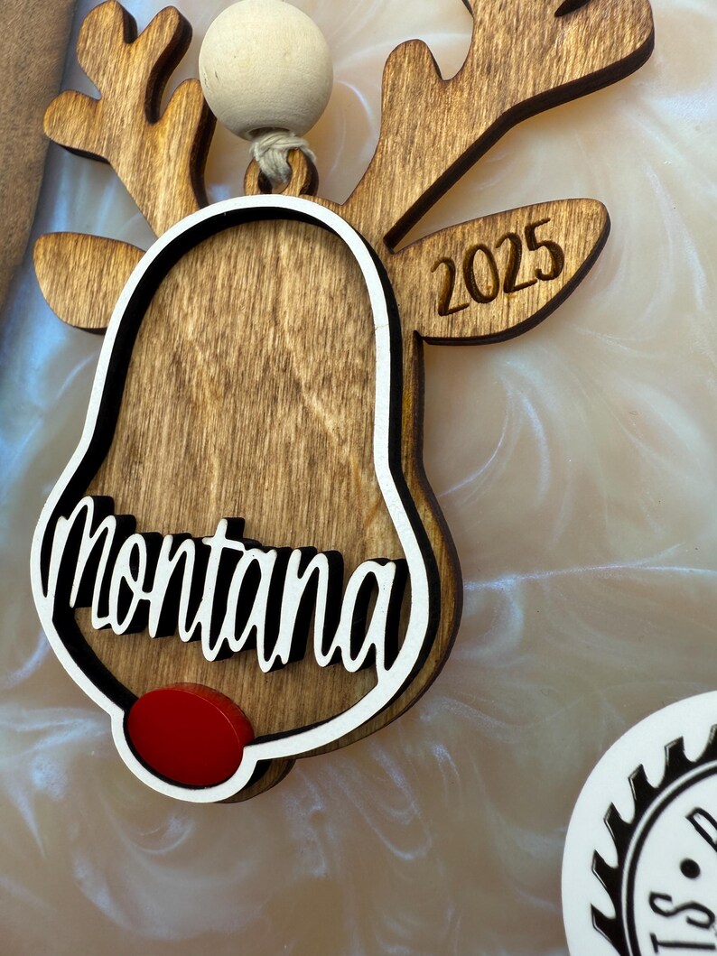 Puede incluir: Adorno de reno de madera con forma de campana y el nombre "Montana" escrito en &eacute;l. El adorno est&aacute; decorado con una nariz roja y el a&ntilde;o "2025" est&aacute; grabado en las astas del reno.