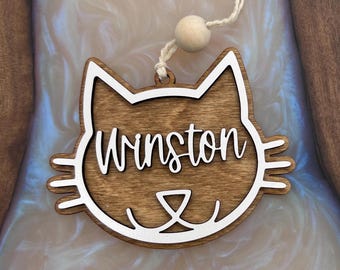 Puede incluir: Un adorno de madera con forma de gato con un contorno blanco y el nombre "Winston" escrito en letras blancas.