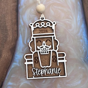 May include: A wooden nutcracker ornament with a white outline. The ornament features a smiling face with a mustache, a crown, and the year 2024. The name Stephanie is engraved on the bottom of the ornament.