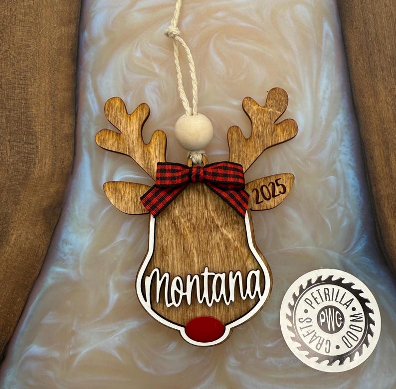 Puede incluir: Adorno de Navidad de reno de madera con un lazo de cuadros rojos y negros, el nombre "Montana" y el a&ntilde;o "2025" grabados en &eacute;l.