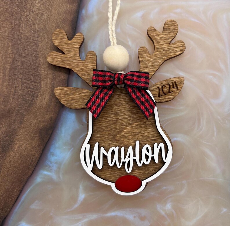 Puede incluir: Adorno de Navidad de reno de madera con un lazo de cuadros rojos y negros, un contorno blanco y el nombre "Waylon" escrito en la parte delantera. El adorno tambi&eacute;n incluye el a&ntilde;o "2024" y una nariz roja.
