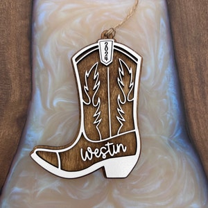 Puede incluir: Un adorno de bota vaquera de madera con un contorno blanco y el nombre "Westin" y el año "2024" grabados en él.